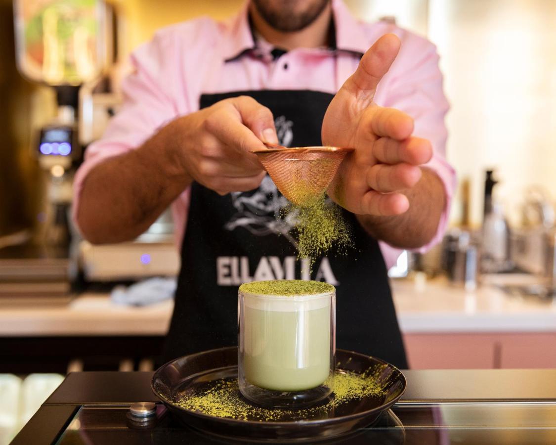 Hotel St Martins Lane Londýn Exteriér fotografie The image shows a person preparing a drink, likely a matcha-based beverage. They are wearing an apron and using a small scoop to sprinkle what appears to be green matcha powder into a glass. The glass is filled with a light green drink, possibly matc
