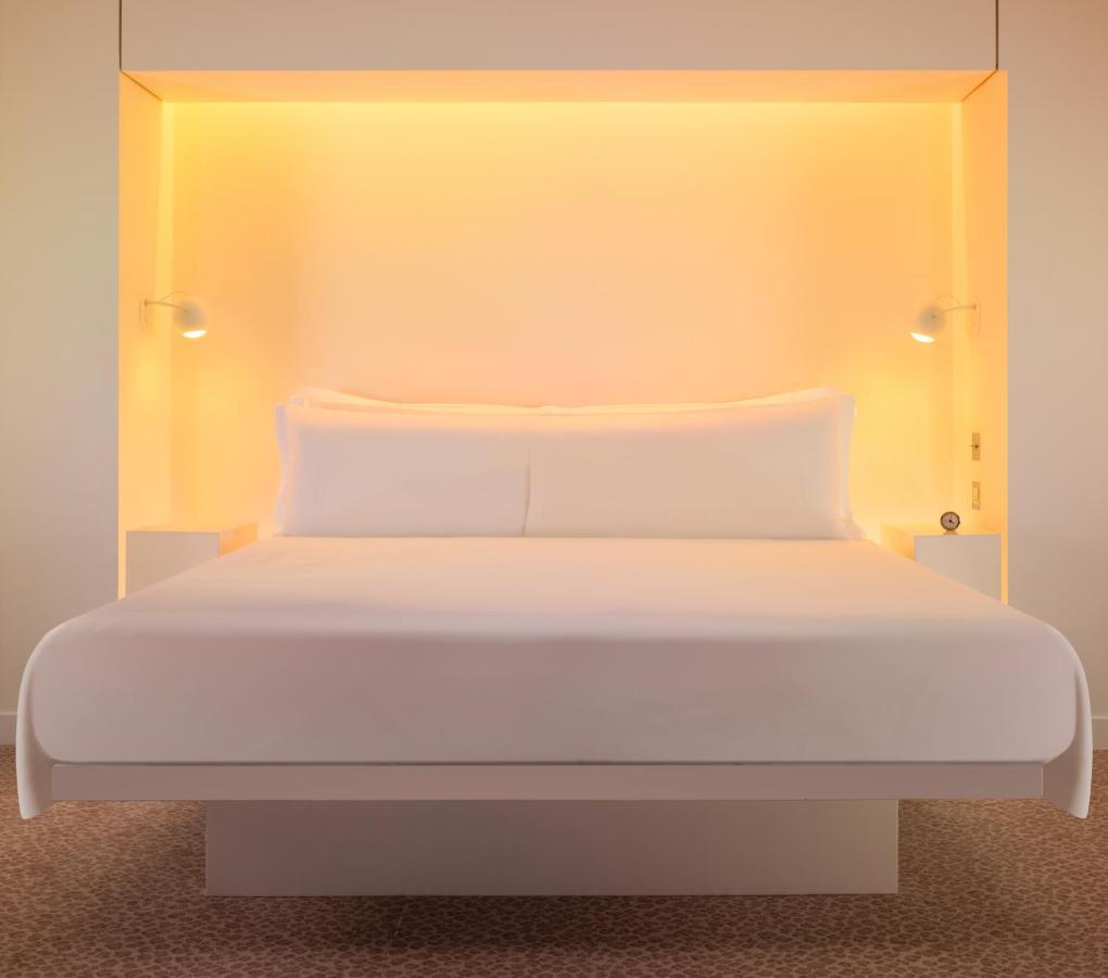 Hotel St Martins Lane Londýn Exteriér fotografie The photo shows a modern and minimalist bedroom setup featuring a large white bed with two pillows. The bed is positioned against a glowing orange backdrop, which adds warmth to the ambiance. On either side of the bed, there are small nightstands equ