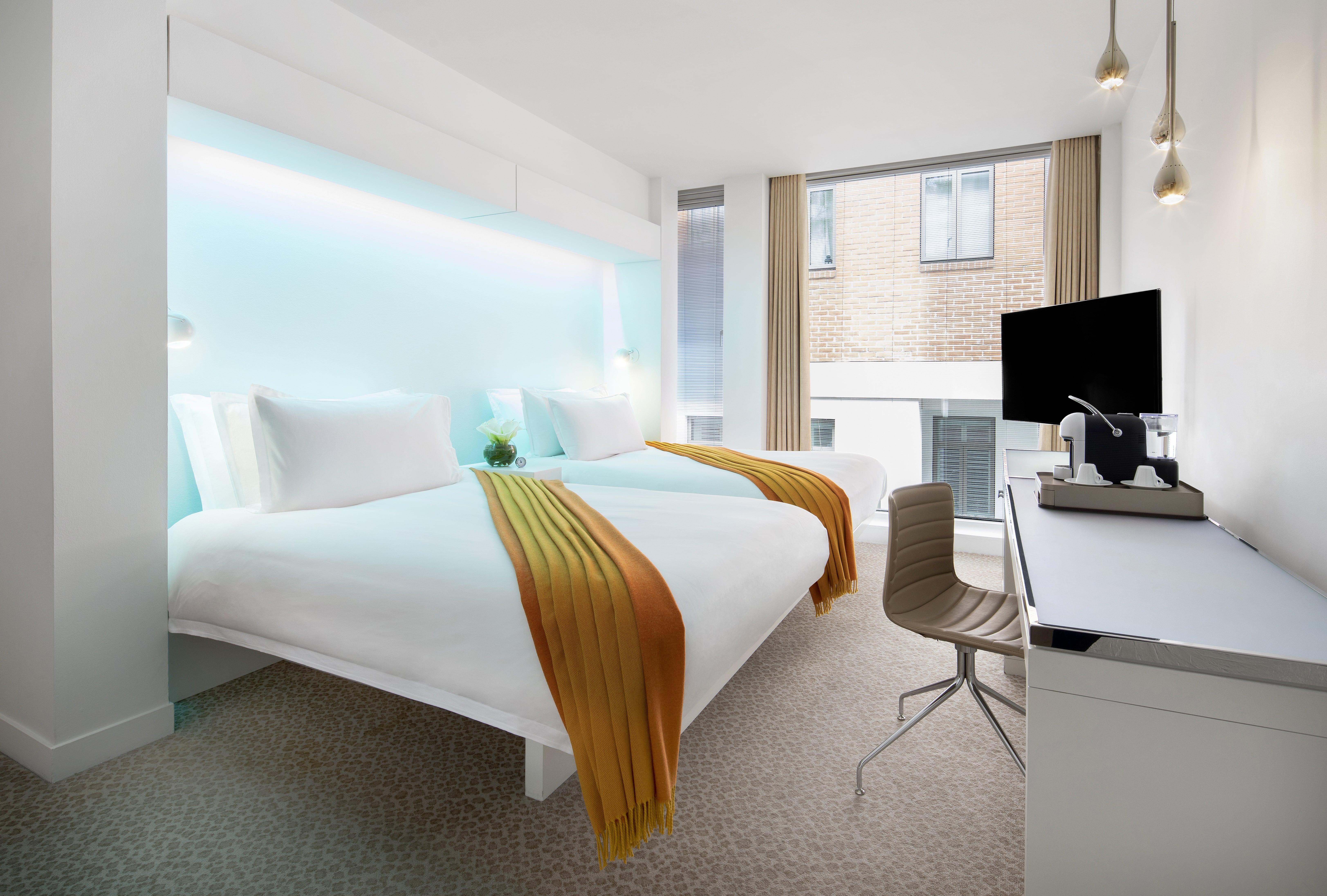 Hotel St Martins Lane Londýn Exteriér fotografie The photo shows a modern hotel room with a clean, contemporary design. There are two beds, each with white bedding and vibrant orange and yellow throws. Behind the beds, there is a glowing blue accent wall, adding a stylish touch. The room features a