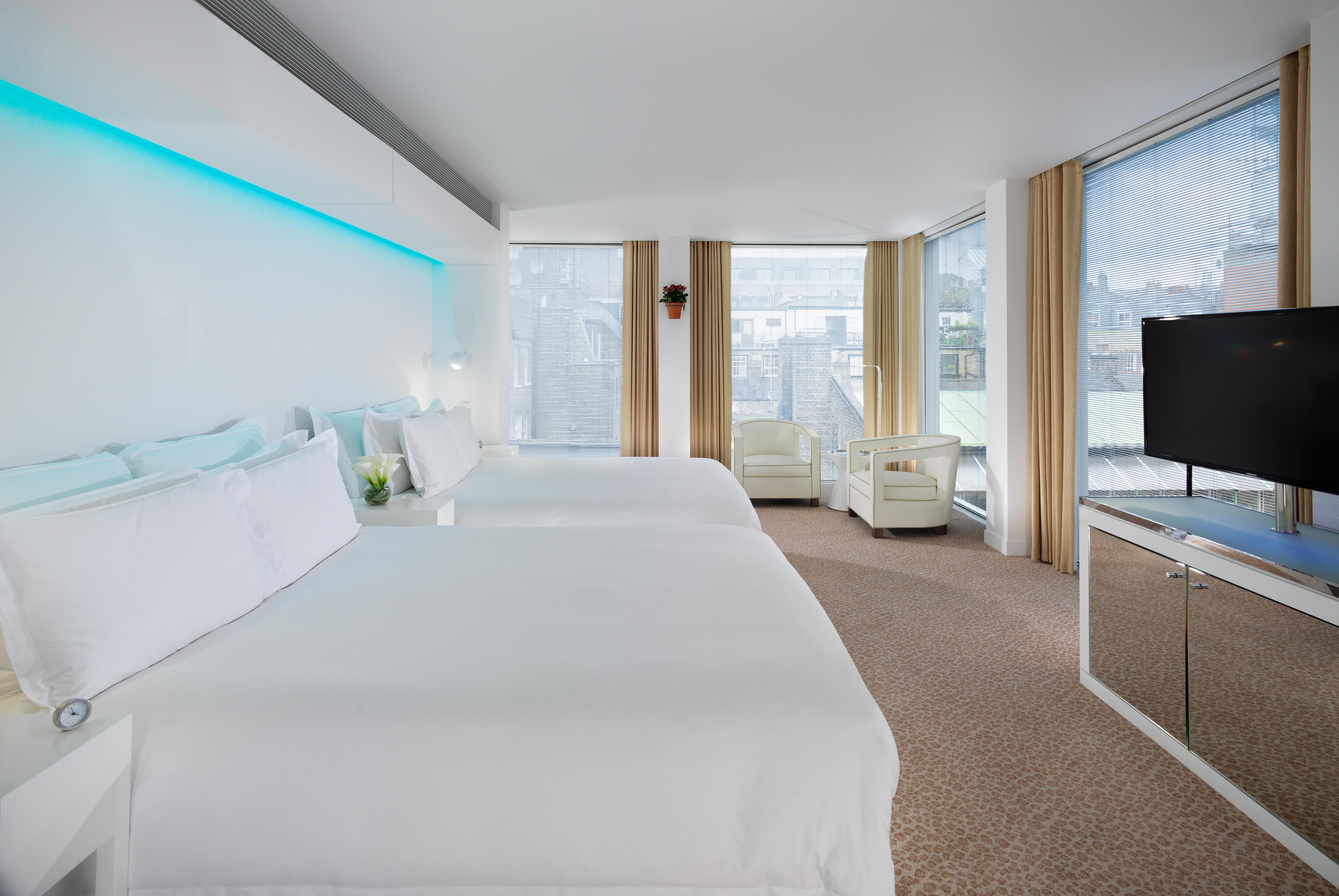 Hotel St Martins Lane Londýn Exteriér fotografie The photo shows a modern hotel room featuring two large beds with white bedding. The room has large windows with light-colored curtains, allowing natural light to fill the space. There is a small seating area with a white sofa and a bedside table. A 