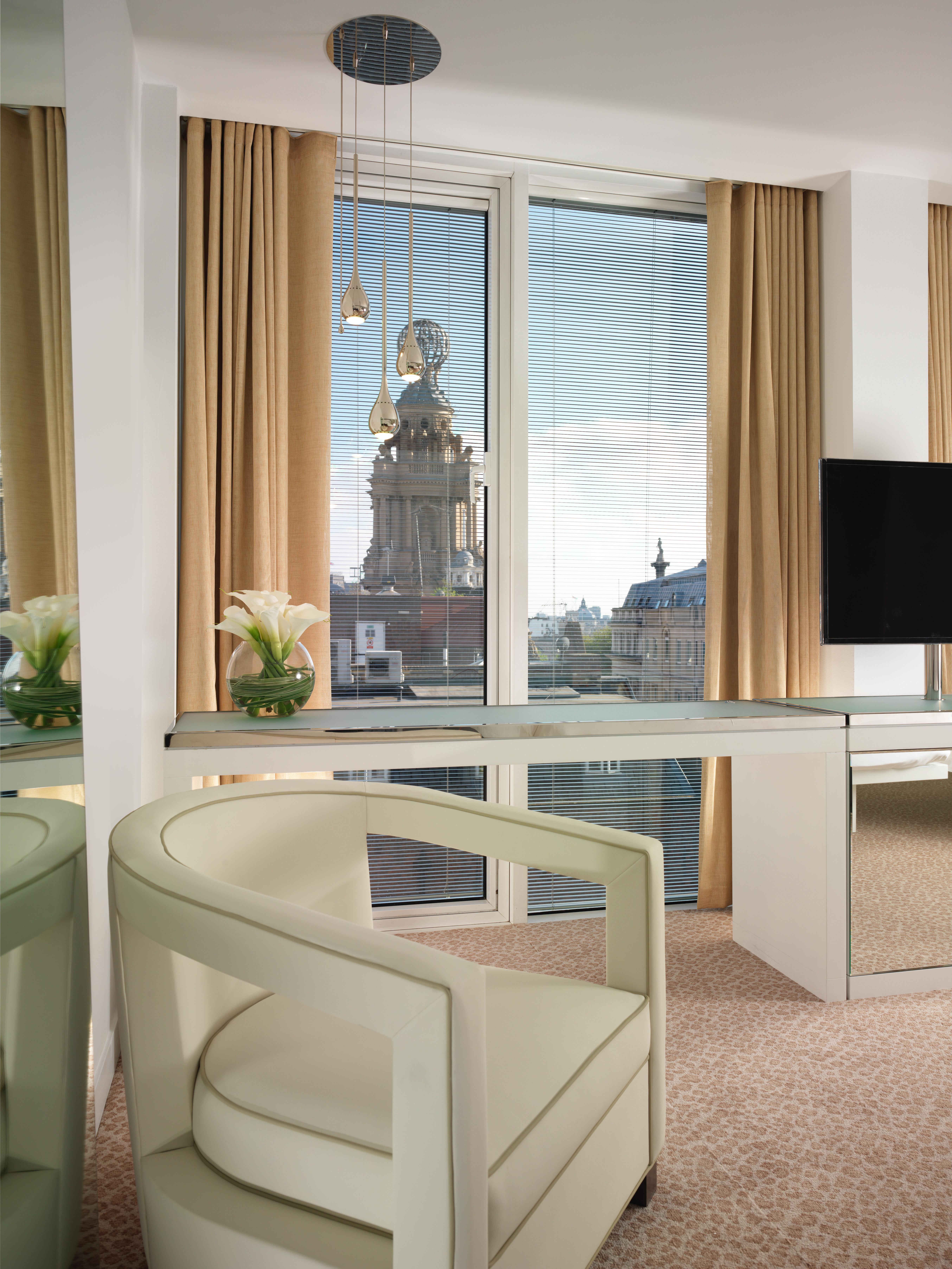 Hotel St Martins Lane Londýn Exteriér fotografie The photo shows a modern interior space, possibly a hotel room or office, featuring a sleek glass desk and a contemporary chair. There’s a vase of white flowers on the desk, and large windows draped with beige curtains provide a view of a building ou