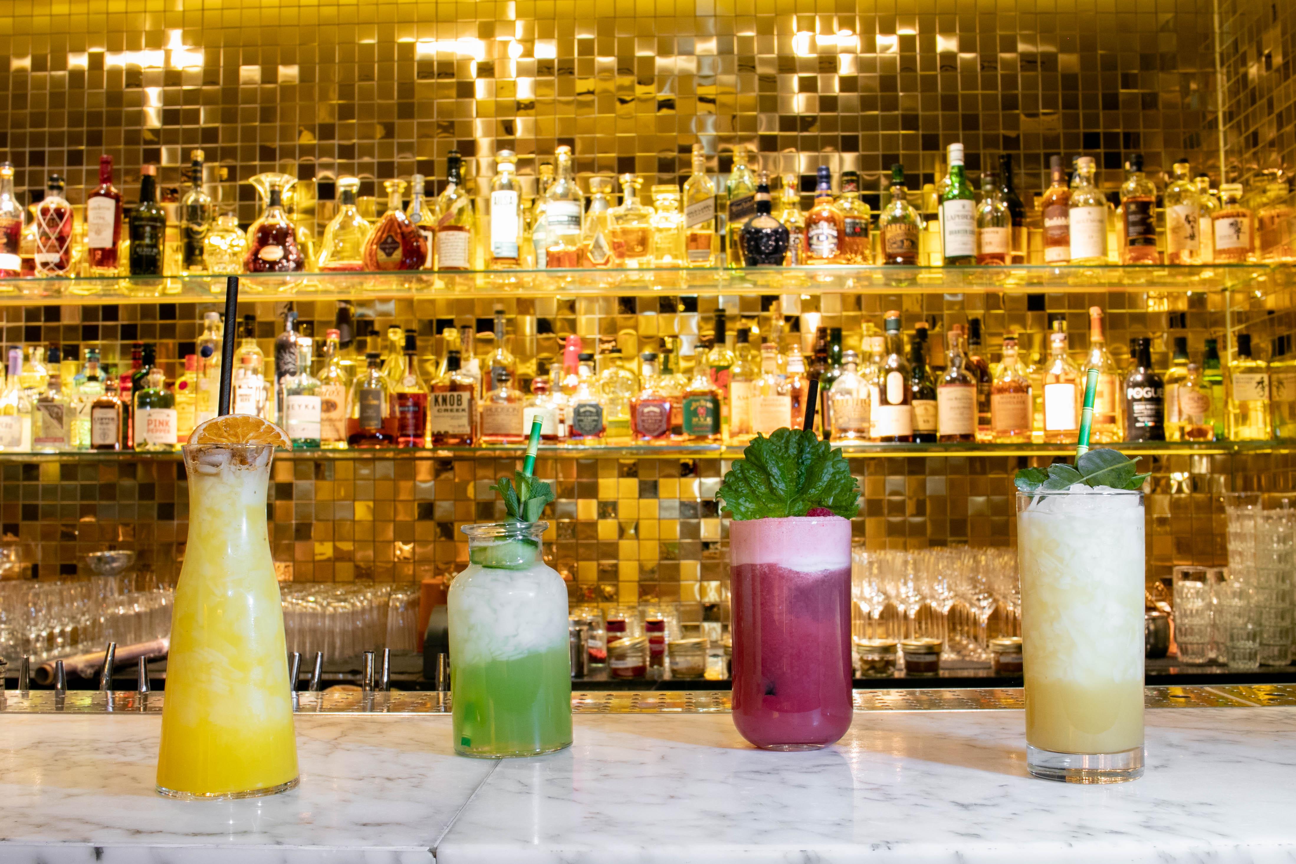 Hotel St Martins Lane Londýn Exteriér fotografie The photo features a stylish bar scene with four colorful cocktails displayed on a marble countertop. Behind the drinks, there is a sparkling, gold mosaic wall lined with various liquor bottles, creating an upscale ambiance. The cocktails include vib