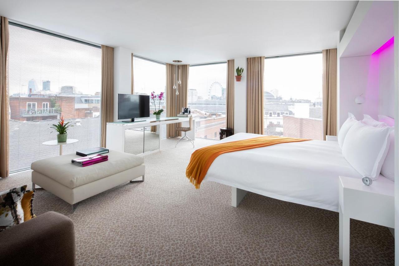 Hotel St Martins Lane Londýn Exteriér fotografie The photo shows a modern hotel room featuring a large bed with white linens and an orange throw blanket. The room has ample natural light coming through large windows with beige curtains, offering a view of a cityscape. There is a small seating area 