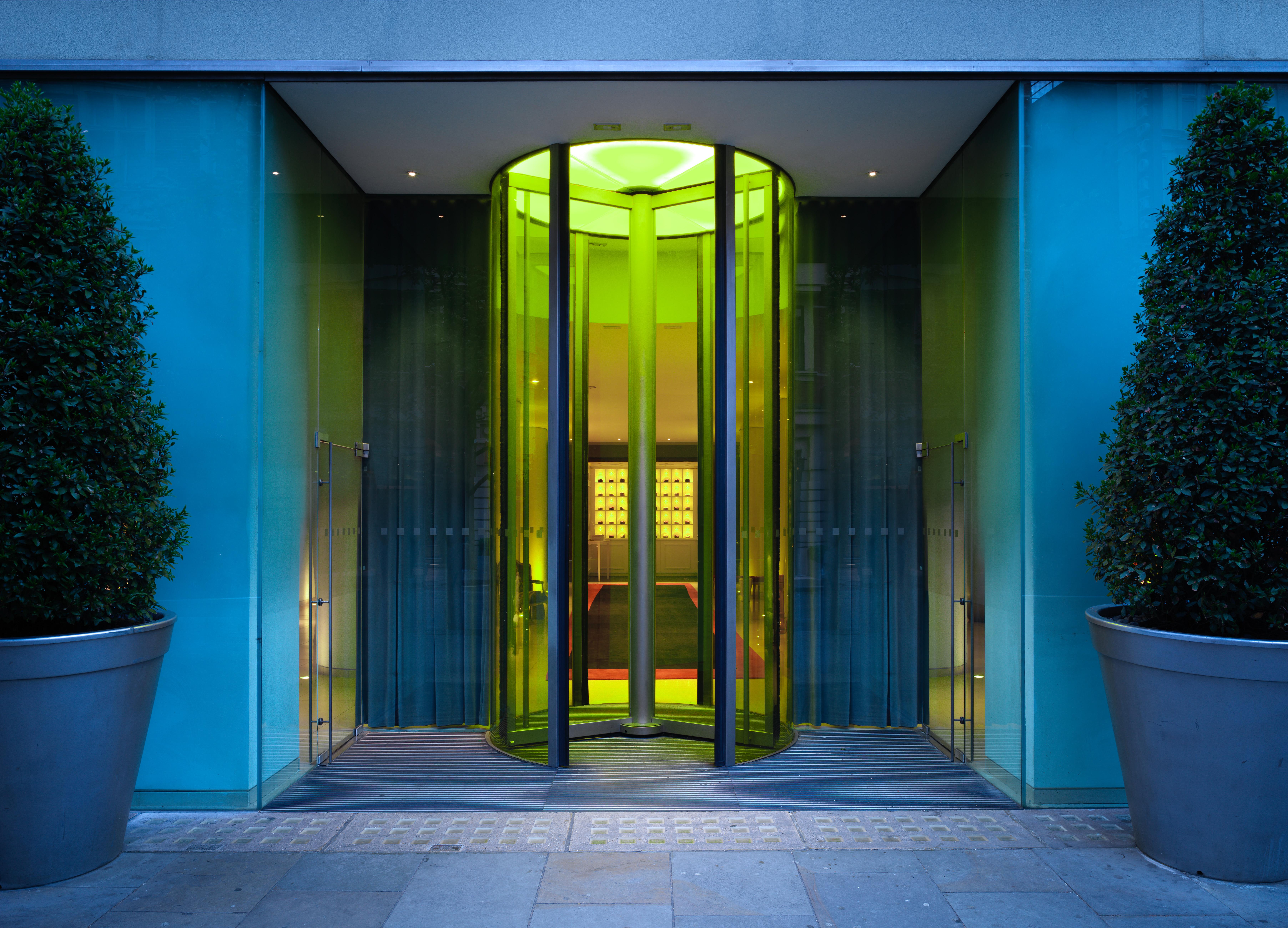 Hotel St Martins Lane Londýn Exteriér fotografie The photo shows a modern glass entrance with a revolving door. The door is surrounded by sleek glass panels that reflect light, creating a vibrant ambiance. The entrance is complemented by large, symmetrical planters containing rounded shrubs on eith