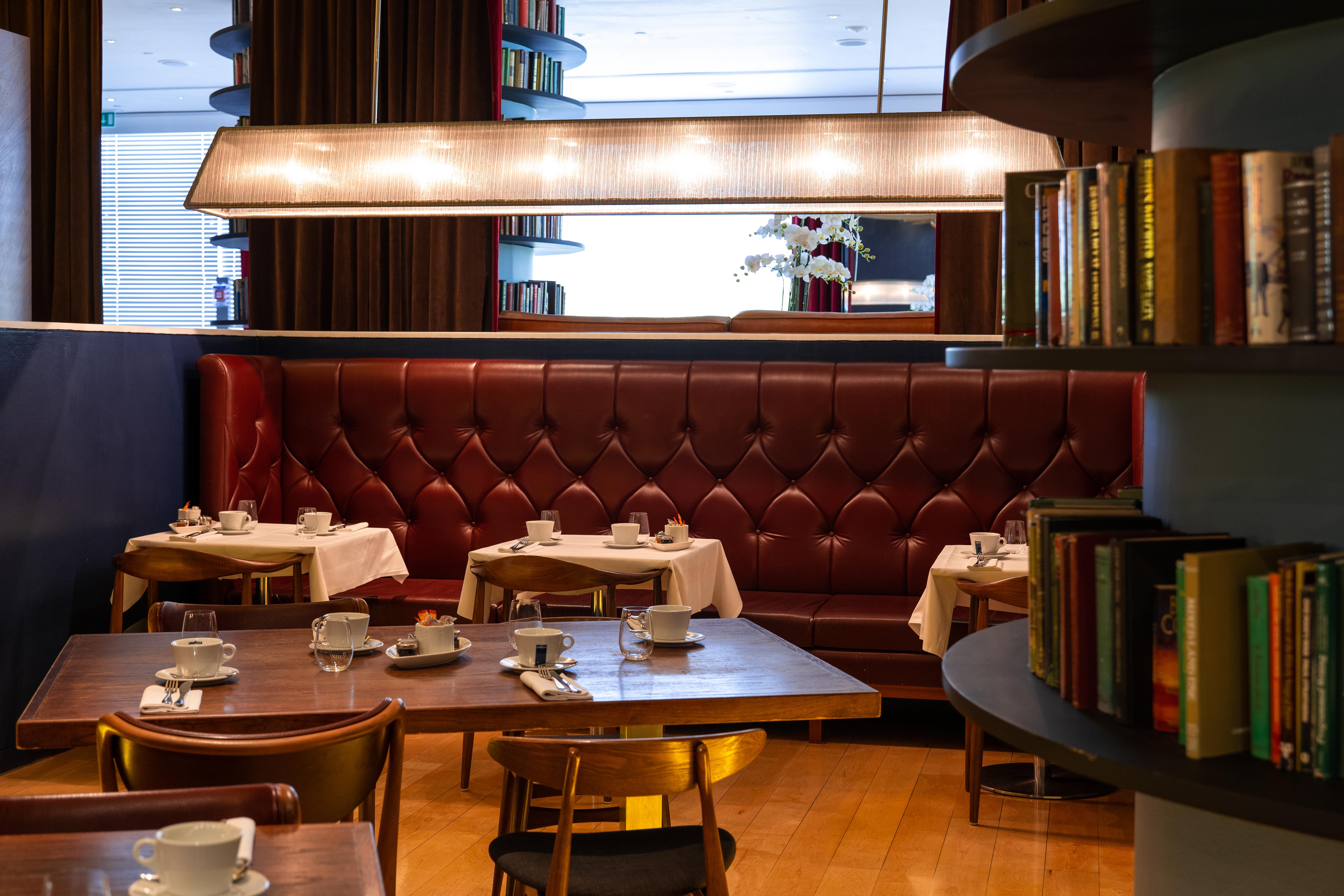 Hotel St Martins Lane Londýn Exteriér fotografie The photo depicts a cozy, elegantly designed restaurant interior. There are several round tables set with white tablecloths and accompanied by small white cups, likely for tea or coffee. The seating includes a plush, burgundy leather banquette agains