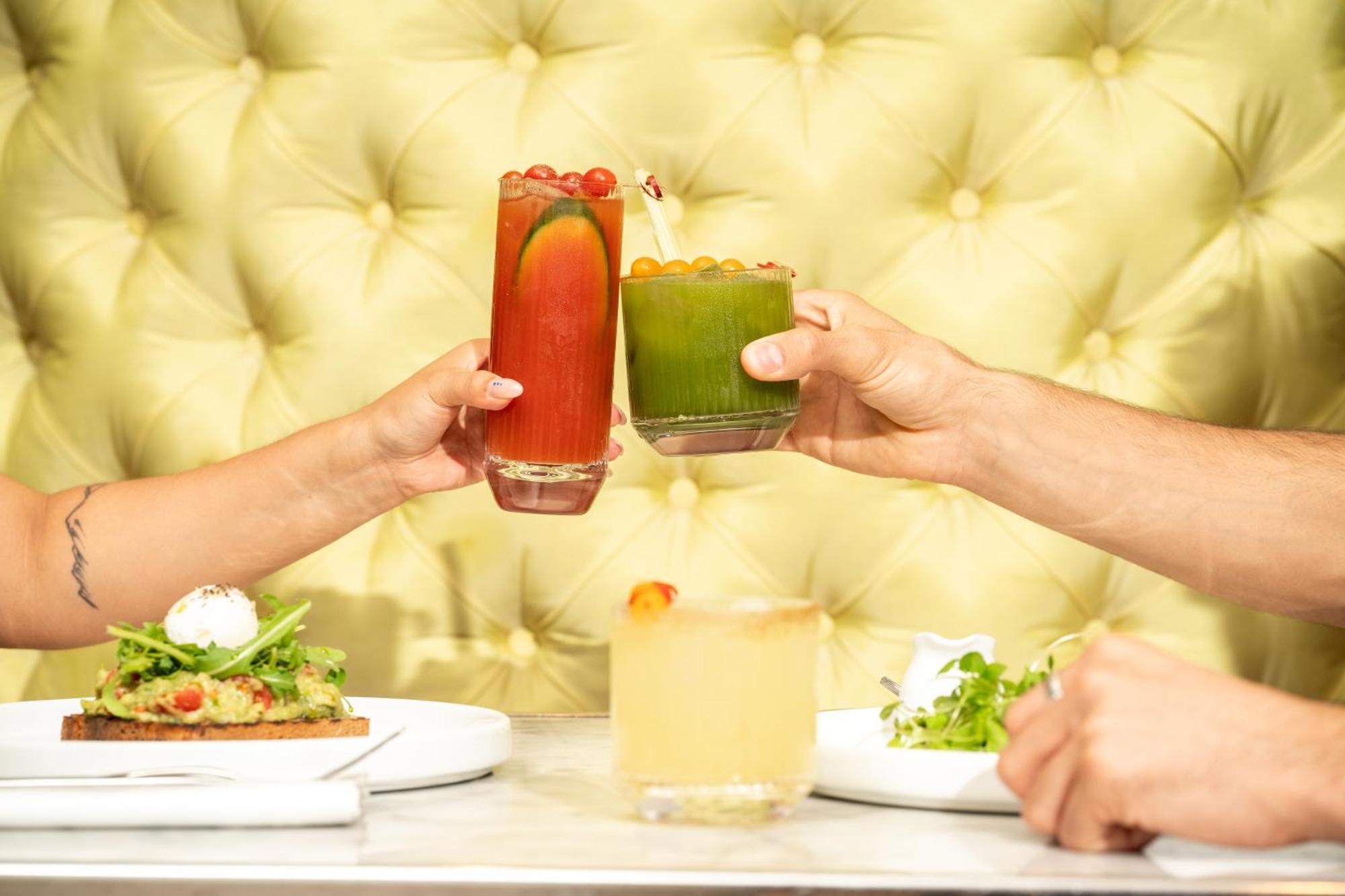 Hotel St Martins Lane Londýn Exteriér fotografie The photo features a vibrant dining scene with two hands clinking glasses in a toast. One glass contains a red drink, possibly a cocktail, adorned with garnishes, while the other is a green beverage, likely a smoothie or juice. In the foreground, the