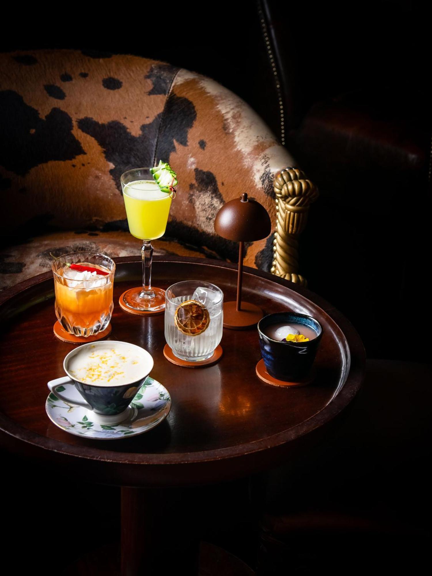 Hotel St Martins Lane Londýn Exteriér fotografie The photo displays a variety of drinks arranged on a round wooden tray. There are five distinct beverages:

1. **A light green cocktail** in a tall glass, garnished with a slice of fruit, suggesting a refreshing flavor.
2. **An orange drink** served 