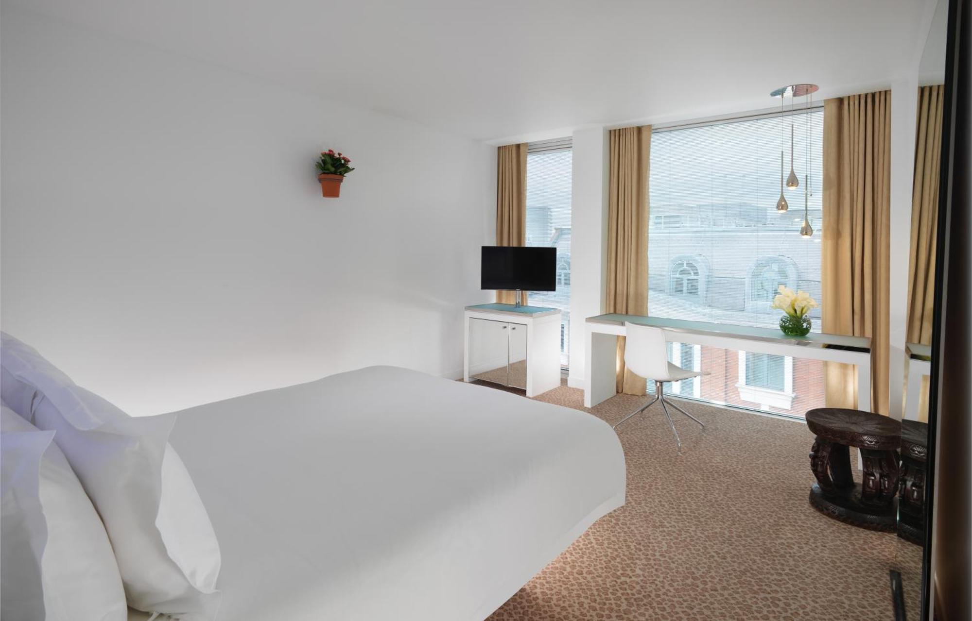 Hotel St Martins Lane Londýn Exteriér fotografie The photo shows a modern hotel room that features a large bed with white linens, a small desk with a chair, and a television mounted on the wall. There are large windows covered with light curtains, allowing natural light to fill the room. A small po